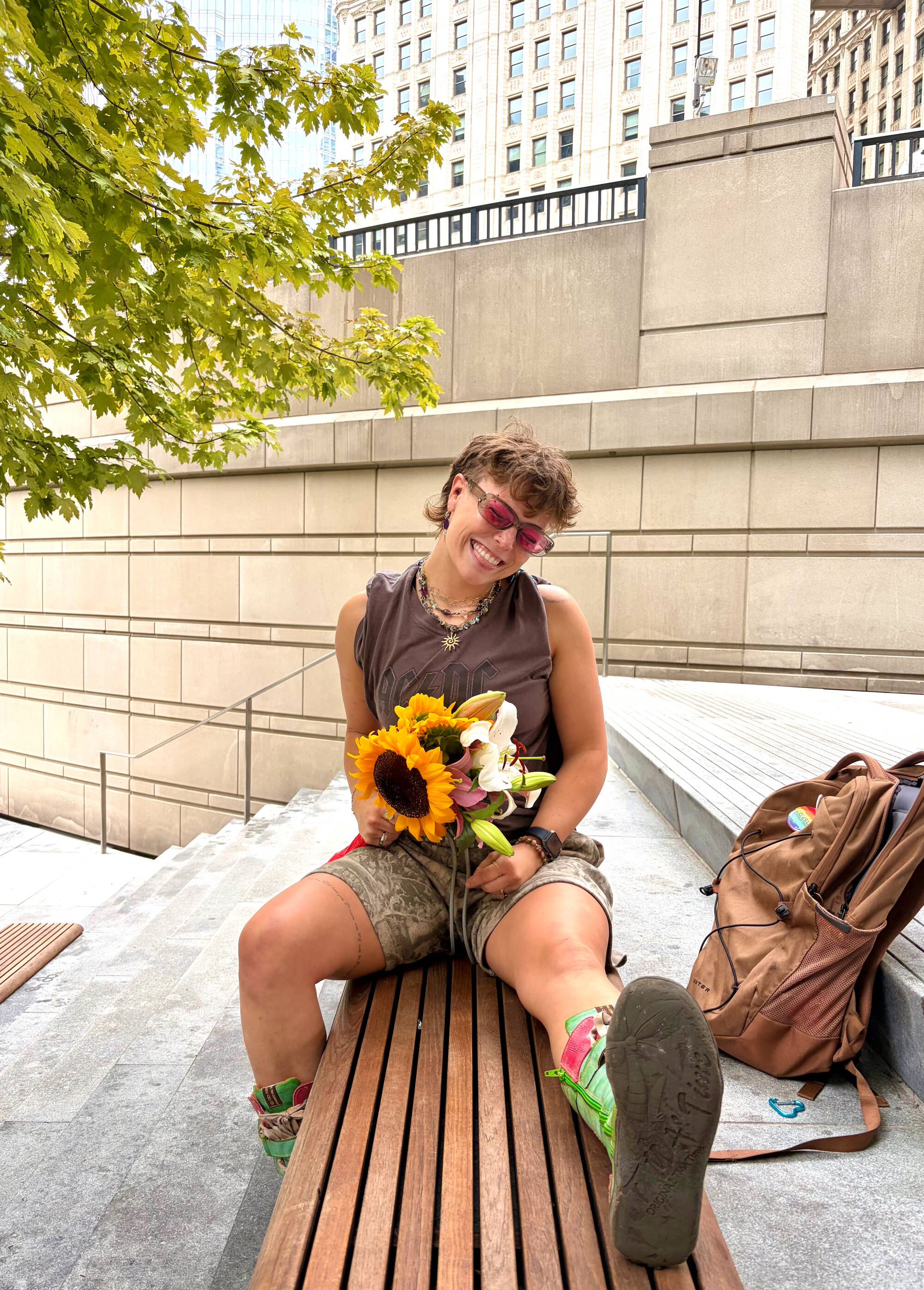 Emily holds a boquet of sunflowers sitting on a wooden bench. She is smiling with her head titled to the side and very cool, tinted sunglasses.